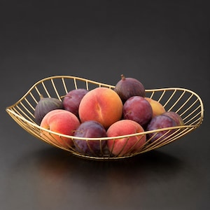 May include: A gold-colored wire fruit bowl with an asymmetrical, wavy rim design. The bowl is filled with a variety of fresh produce, including two fuzzy orange and pink peaches, several dark purple plums, and two dark-skinned, teardrop-shaped figs.