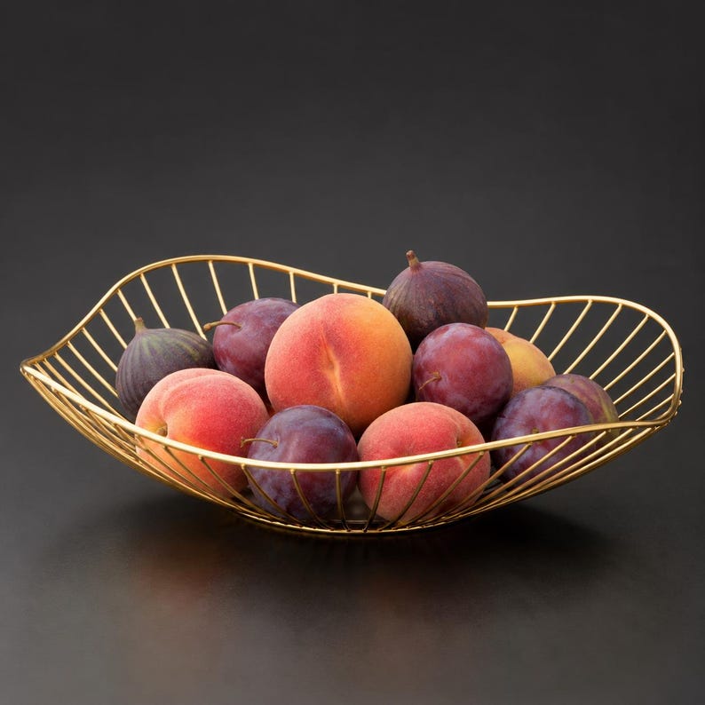May include: A gold-colored wire fruit bowl with an asymmetrical, wavy rim design. The bowl is filled with a variety of fresh produce, including two fuzzy orange and pink peaches, several dark purple plums, and two dark-skinned, teardrop-shaped figs.