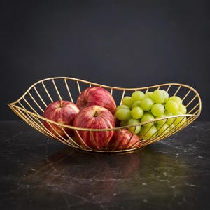 May include: A gold-colored wire fruit bowl with an elongated, curved shape containing three red apples and a large cluster of green grapes. The bowl is constructed from thin metal rods arranged in a symmetrical, open-frame design.