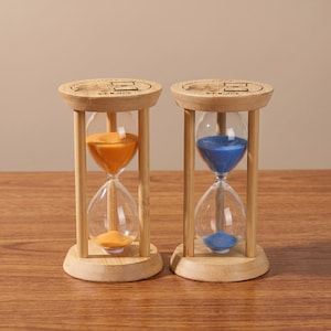 May include: Two hourglasses with wooden frames and bases. One contains orange sand, the other blue. The clear glass bulbs are held by vertical wooden supports. The top and bottom of each hourglass are circular, with etched designs.