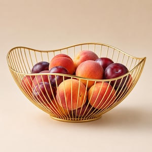 May include: A gold-colored wire fruit bowl with a wavy, open-frame design filled with a variety of fresh peaches and dark purple plums. The bowl features a circular base and thin, vertical metal rods that curve upward to form the basket shape.