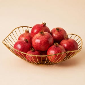 May include: A gold-toned wire fruit bowl with a wavy, open-frame design holding eight whole pomegranates. The pomegranates are round with deep red skins and textured crowns, arranged in a cluster within the metallic, curved wire structure.