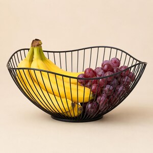 May include: A black wire fruit basket with a wavy, open-frame design holding a bunch of yellow bananas and a cluster of red grapes. The basket features a circular base and thin metal rods that curve upward to form a wide, bowl-like structure.