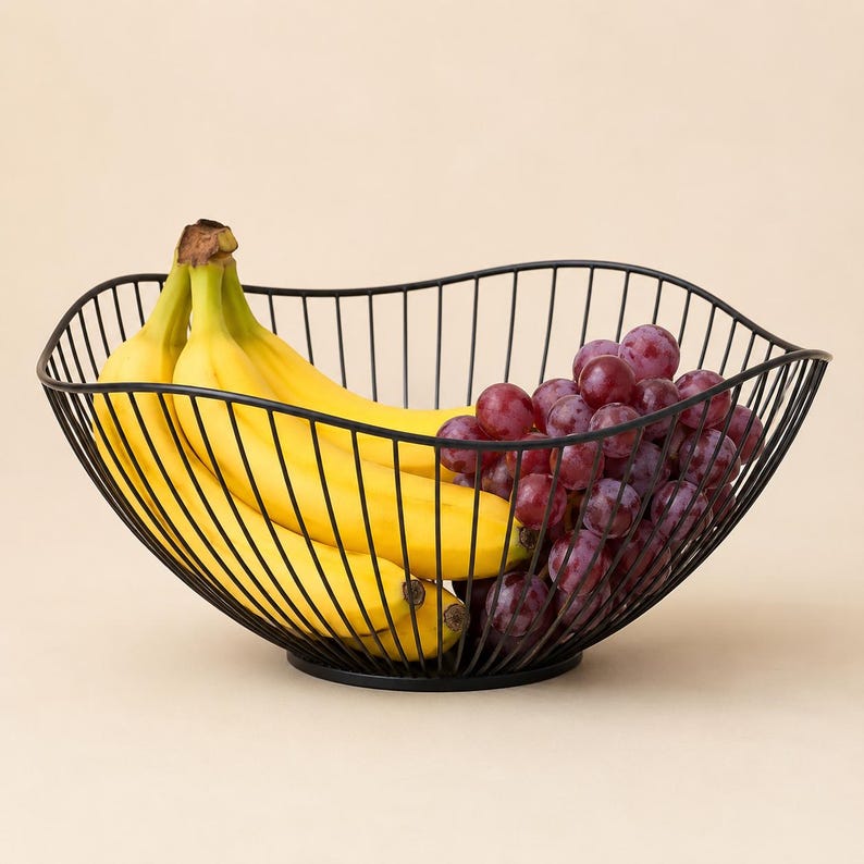 May include: A black wire fruit basket with a wavy, open-frame design holding a bunch of yellow bananas and a cluster of red grapes. The basket features a circular base and thin metal rods that curve upward to form a wide, bowl-like structure.