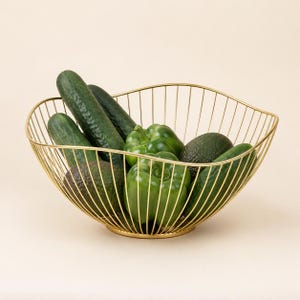 May include: A gold-colored wire fruit basket with a curved, open-frame design. The basket is filled with fresh green produce, including two dark green cucumbers, two green bell peppers, and two dark green avocados, all resting inside the metal frame.