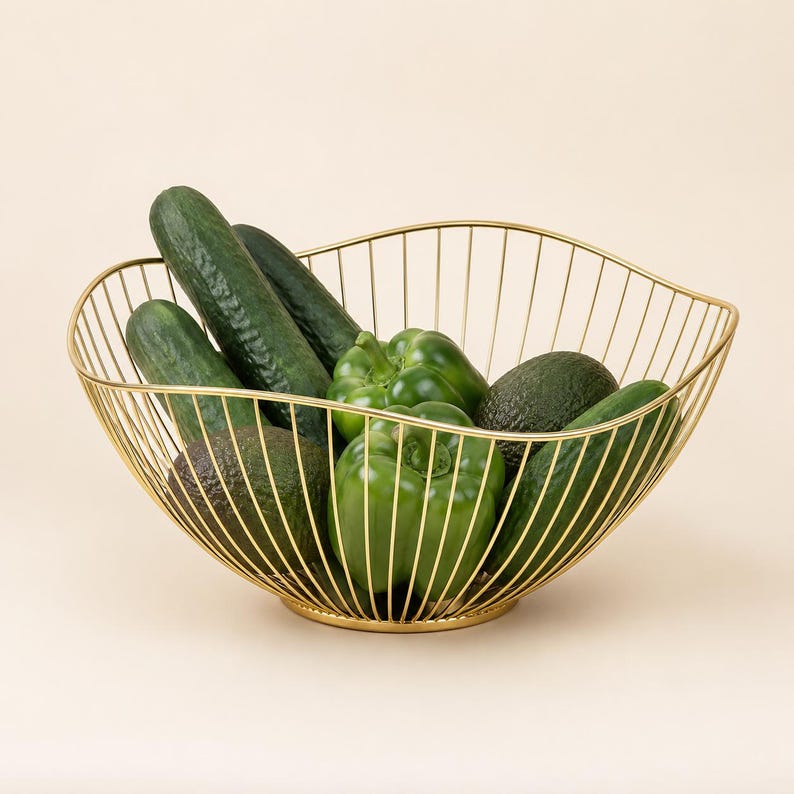 May include: A gold-colored wire fruit basket with a curved, open-frame design. The basket is filled with fresh green produce, including two dark green cucumbers, two green bell peppers, and two dark green avocados, all resting inside the metal frame.