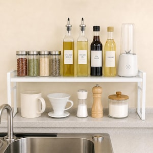 May include: A white metal kitchen shelf holding four spice jars, two tall oil bottles labeled Extra Virgin Olive Oil and Avocado Oil, a soy sauce bottle, a vinegar bottle, and a portable blender. Below are a mug, a coffee dripper, a salt shaker, a pepper mill, and a jar.