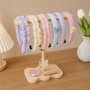 Op de afbeelding: Een licht houten displaystandaard met zes haarbanden in diverse kleuren en texturen, waaronder geplooide, effen en gingham patronen. De standaard heeft een bloemvormig accent, een klein rond bakje en een houten haarclip op de voet.