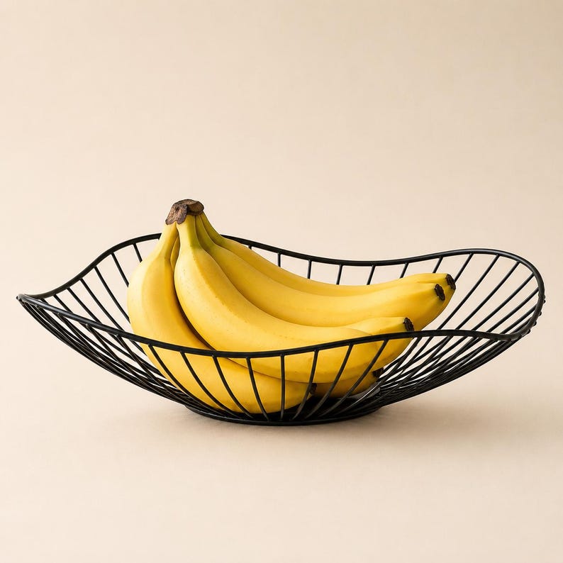 May include: A black wire fruit bowl with a wavy, open-frame design holding a bunch of bright yellow bananas. The bowl features a curved, elongated shape constructed from thin metal rods, providing a clear view of the fruit resting inside.