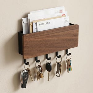 May include: A wall-mounted key holder and mail organizer. It features a dark wood front with a black metal frame and hooks. The organizer holds mail and postcards, with keys hanging from the hooks below. The keys include a car key fob and other keys.