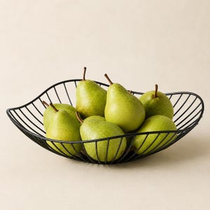 May include: A black wire fruit bowl with a curved, open-frame design holding six fresh green pears. The bowl features a series of thin, parallel metal rods that form a wide, shallow basin, providing a clear view of the fruit inside.