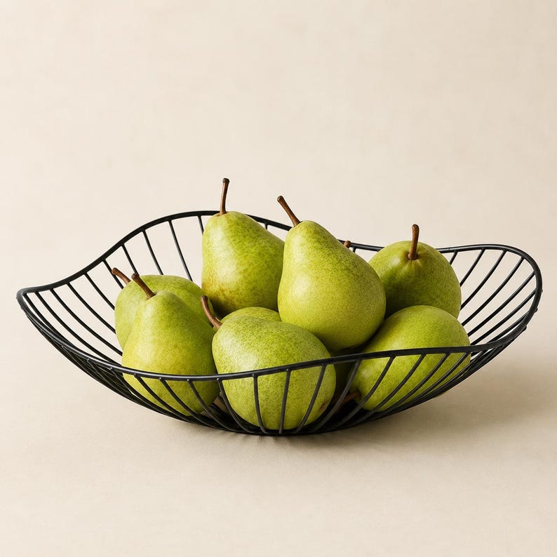 May include: A black wire fruit bowl with a curved, open-frame design holding six fresh green pears. The bowl features a series of thin, parallel metal rods that form a wide, shallow basin, providing a clear view of the fruit inside.
