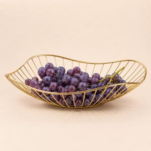 May include: A gold-colored wire fruit bowl with an elongated, wavy-edged design, filled with a cluster of fresh, dark purple grapes. The bowl is constructed from thin, vertical metal rods that create an open, airy, and cage-like structure.