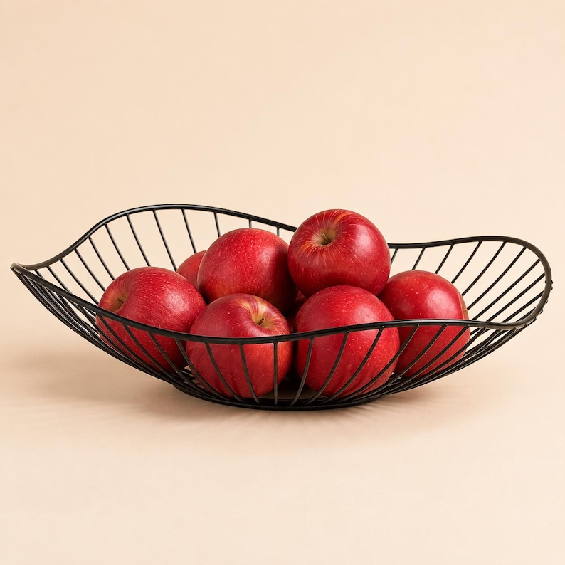 May include: A black wire fruit bowl with a curved, open-frame design holding several bright red apples. The bowl features a series of thin, parallel metal rods that form a wide, shallow basin shape, containing the round, smooth-skinned red fruit.