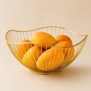May include: A gold-colored wire fruit bowl with a wavy, open-frame design containing five ripe, yellow-orange mangoes. The bowl features a circular base and thin, vertical metal rods that curve upward to form a wide, scalloped rim.