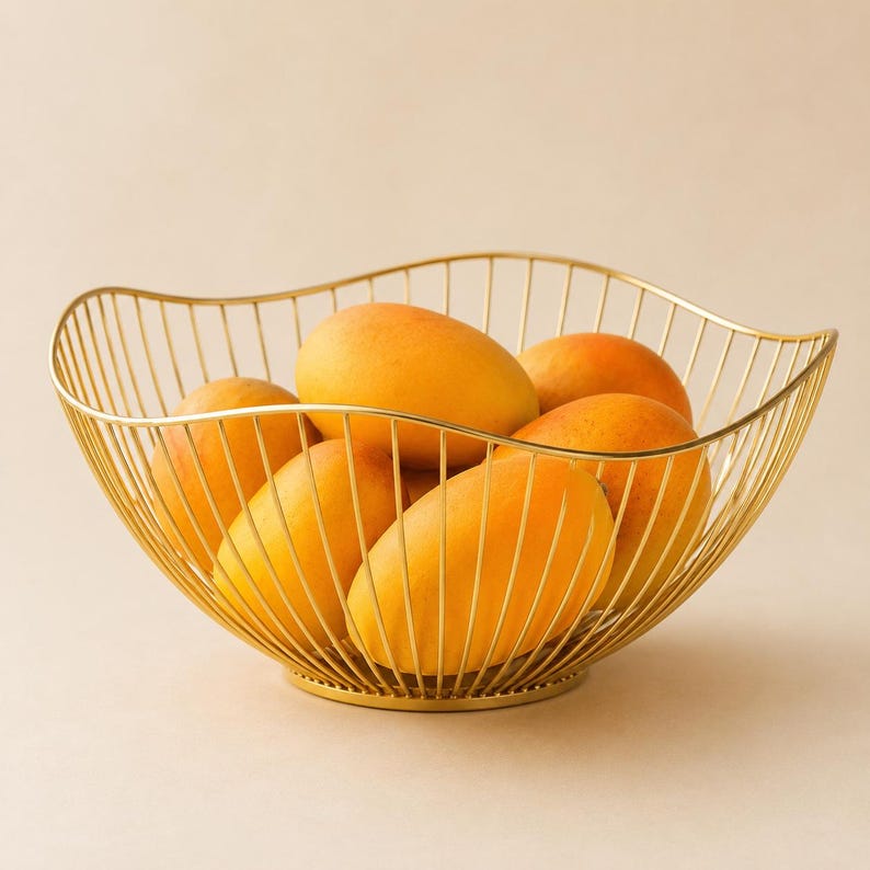May include: A gold-colored wire fruit bowl with a wavy, open-frame design containing five ripe, yellow-orange mangoes. The bowl features a circular base and thin, vertical metal rods that curve upward to form a wide, scalloped rim.
