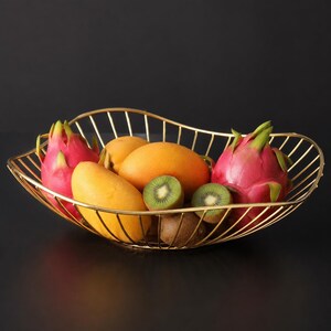 May include: A gold-toned wire fruit basket with an oval, wavy-edged design. The basket contains two bright pink dragon fruits, two smooth yellow mangoes, two brown kiwis, and two sliced green kiwi halves with visible black seeds inside the wire frame.