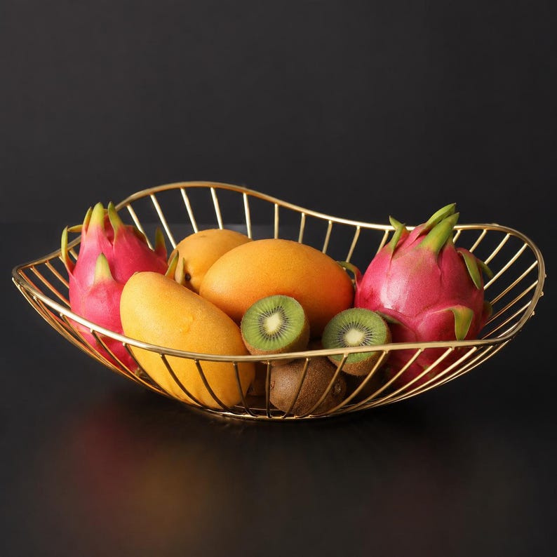 May include: A gold-toned wire fruit basket with an oval, wavy-edged design. The basket contains two bright pink dragon fruits, two smooth yellow mangoes, two brown kiwis, and two sliced green kiwi halves with visible black seeds inside the wire frame.