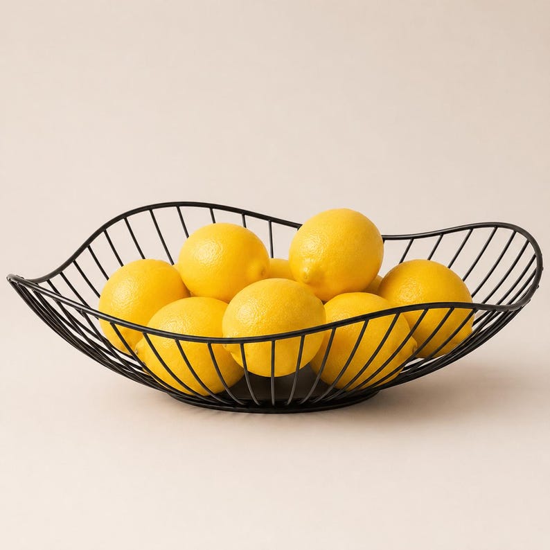 May include: A black wire fruit bowl with a wavy, leaf-like rim design, filled with several bright yellow lemons. The bowl is constructed from thin, evenly spaced metal rods that curve upward to form an open, airy structure for holding fruit.