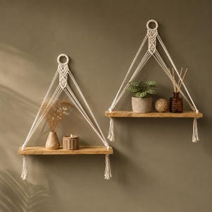 May include: Two wall-mounted floating shelves made of light-toned wood, each suspended by cream-colored macrame cord featuring intricate knotting and a circular wooden ring at the top. The shelves hold a small potted plant, a candle, and a vase.