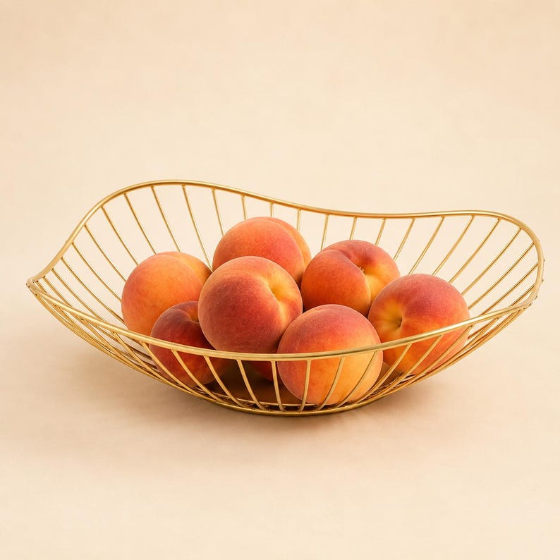 May include: A gold-colored wire fruit bowl with an asymmetrical, curved rim design. The bowl is filled with six fresh, round peaches that have a gradient of yellow, orange, and red skin tones. The wire structure is thin and open-framed.