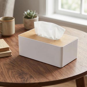 Puede incluir: Un dispensador de pañuelos rectangular con una base de plástico blanco sólido y una tapa de madera de color marrón claro con una abertura central. Un pañuelo de papel blanco sobresale por la abertura superior de la caja.