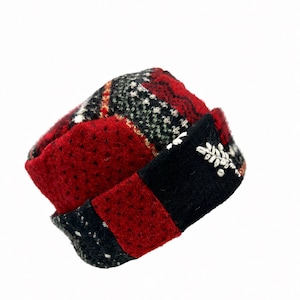 May include: A patchwork hat constructed from various wool-like fabric segments in shades of red, black, and white. The design features a folded brim with a small, white embroidered leaf-like motif on the front panel and a textured surface.