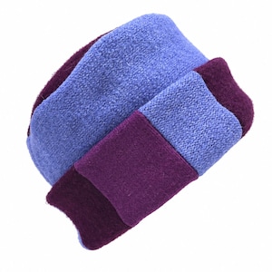 May include: A knit hat featuring a color-blocked design with alternating sections of textured periwinkle blue and solid deep plum purple yarn. The hat has a rounded crown and a wide, folded brim that wraps around the base in a layered pattern.