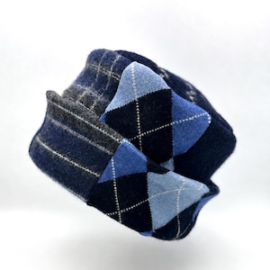 May include: A knit hat featuring a combination of patterns, including a dark blue and light blue argyle design on the brim and a dark blue and grey plaid pattern on the crown. The hat is constructed from a textured, soft-looking knit fabric.