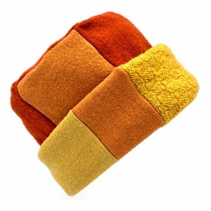 May include: A knit beanie featuring a color-blocked design with panels in shades of burnt orange, pumpkin orange, mustard yellow, and a textured cable-knit section in bright yellow. The hat is constructed from multiple distinct fabric segments.