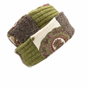 May include: A patchwork headband constructed from various knit fabric segments. The design features olive green ribbed sections, dark brown textured panels, and a cream-colored piece with a circular floral pattern in shades of green and pink.