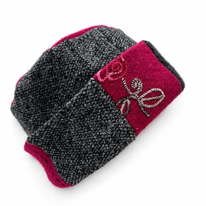 May include: A folded beanie hat featuring a dark charcoal grey knit texture contrasted with a vibrant magenta felt band. The magenta section is decorated with intricate, swirling silver and pink thread embroidery in a floral-inspired pattern.