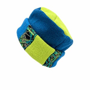 May include: A structured hat featuring alternating panels of deep blue and bright lime green felted fabric. The design includes a wide band of lime green felt and two decorative sections of intricate, multi-colored woven yarn trim.