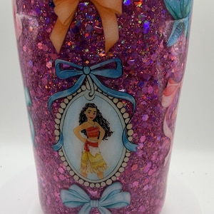 May include: A cylindrical tumbler covered in pink glitter with a glossy finish. The design features a central oval portrait of a character with long dark hair wearing a red and tan outfit, framed by blue ribbons and bows, with additional orange bows.