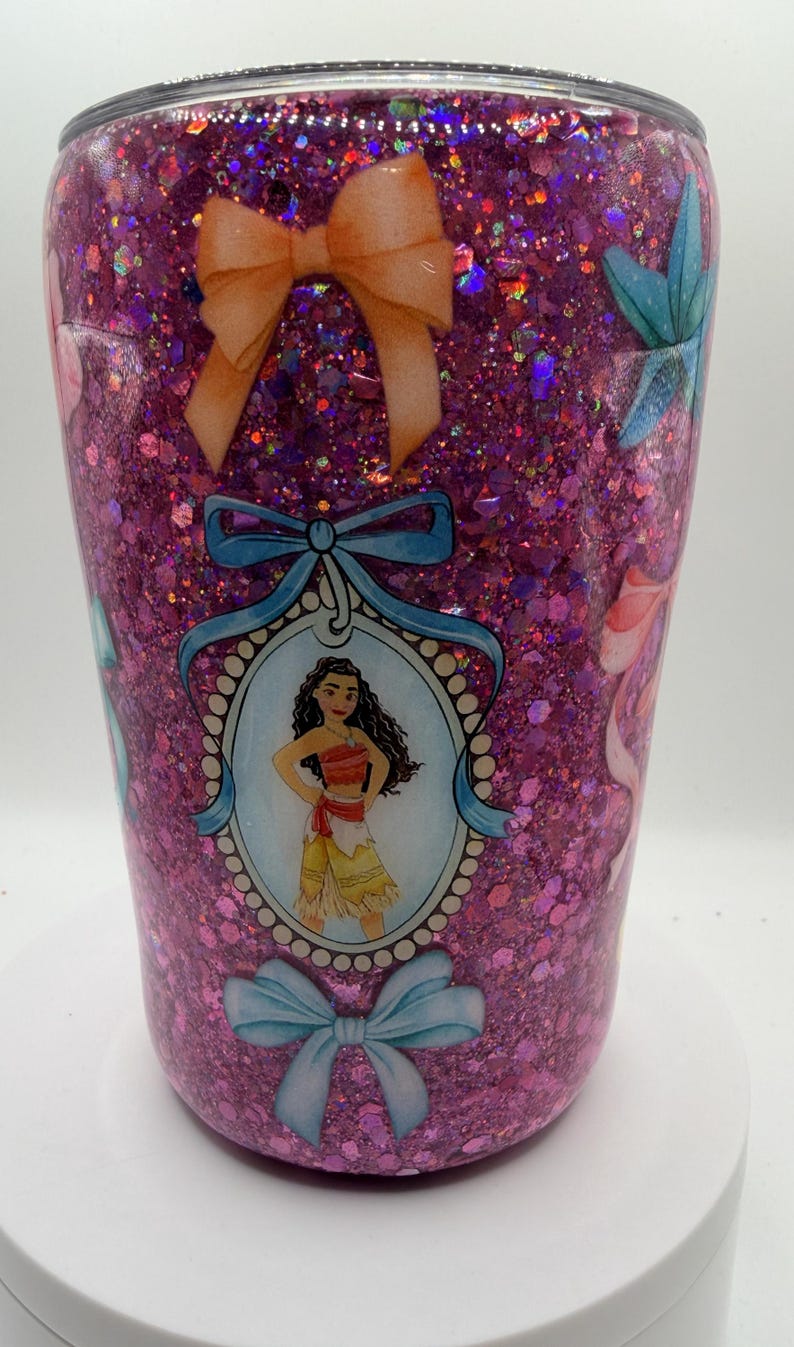 May include: A cylindrical tumbler covered in pink glitter with a glossy finish. The design features a central oval portrait of a character with long dark hair wearing a red and tan outfit, framed by blue ribbons and bows, with additional orange bows.