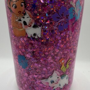 May include: A pink glitter tumbler featuring a clear lid and various character decals. The surface is covered in dense, iridescent pink glitter. Decals include a small child, a white piglet, a rooster, and several blue and purple hibiscus flowers.