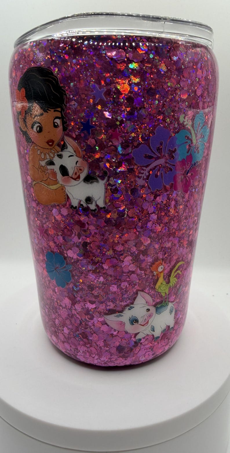 May include: A pink glitter tumbler featuring a clear lid and various character decals. The surface is covered in dense, iridescent pink glitter. Decals include a small child, a white piglet, a rooster, and several blue and purple hibiscus flowers.