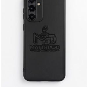 Può includere: Una custodia per smartphone nera opaca con un ritaglio per tripla fotocamera nell'angolo in alto a sinistra e una piccola apertura circolare per il flash. Il centro presenta un logo in rilievo con il testo MATTRIX3D e un'icona grafica stilizzata.