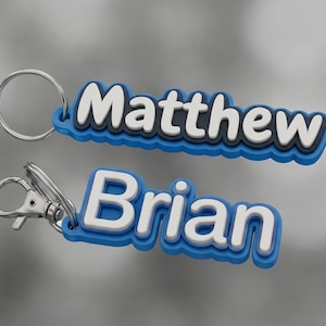 Puede incluir: Dos llaveros personalizados con los nombres "Matthew" y "Brian" en blanco, contorneados en negro, sobre un fondo azul. Cada llavero tiene un llavero o mosquetón plateado.