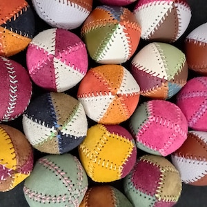 May include: A collection of multi-colored juggling balls constructed from stitched leather panels. Each ball features a geometric pattern of various colors including pink, orange, yellow, green, brown, and white, joined by visible white stitching.