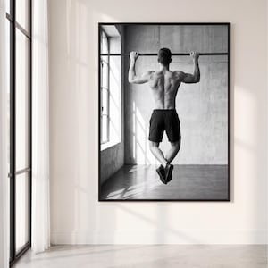 Könnte beinhalten: Ein gerahmter schwarz-weiß Wandkunstdruck, der eine Person bei Klimmzügen an einer horizontalen Stange zeigt. Die Person ist von hinten zu sehen, trägt schwarze Sportshorts und dunkle Turnschuhe vor einer Betonwand als Hintergrund.