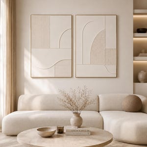 Könnte beinhalten: Zwei gerahmte abstrakte Wandkunstwerke in Beige- und Goldtönen. Das Kunstwerk zeigt geometrische Formen und Linien. Die Stücke werden über einem beigen Sofa und einem runden Couchtisch mit einer Vase mit getrockneten Blumen ausgestellt.