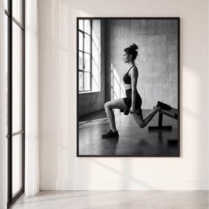 Pode incluir: Uma impressão de arte de parede emoldurada em preto e branco retratando uma pessoa realizando um exercício de agachamento búlgaro. O indivíduo veste um top esportivo e shorts, segurando um haltere em cada mão sobre um banco de exercícios.
