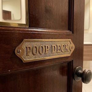 Könnte beinhalten: Eine dunkelbraune Holztür mit einem Messingschild mit der Aufschrift "POOP DECK". Das Schild ist rechteckig mit abgerundeten Kanten und mit Schrauben an der Tür befestigt. Ein runder, dunkler Metalltürknauf ist sichtbar.
