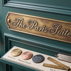 May include: A dark teal door featuring a rectangular brass plaque engraved with The Paris Suite, a brass key with a matching tag, and a display shelf holding circular metal signs labelled Room 101, Suite A, and a small ruler for scale.