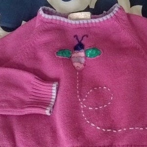 May include: A pink sweater with a bee applique. The sweater features a ribbed hem and cuffs with a light purple trim. The bee is embroidered with purple, green, and pink details, and a dotted line trails behind it.