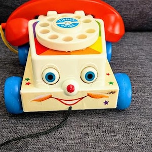 May include: A vintage Fisher-Price toy phone with a smiling face, blue eyes, and a red nose. The toy has a red handset, a rotary dial, and blue wheels. The phone is cream-colored with colorful accents and a black cord.