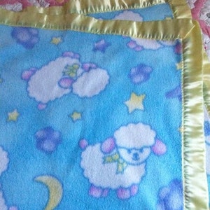 May include: A light blue fleece blanket featuring a repeating pattern of white sheep with pink ears, yellow stars, purple clouds, and yellow crescent moons. The blanket is finished with a shiny, pale yellow satin trim along the edges.