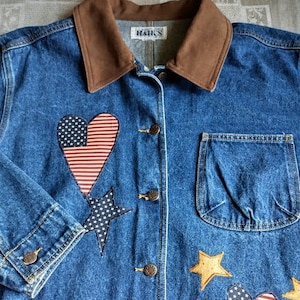 May include: A blue denim jacket with a brown corduroy collar. The jacket has several patches sewn on, including hearts, stars, and a star-spangled banner design. The text "Hanks" is visible on the label.