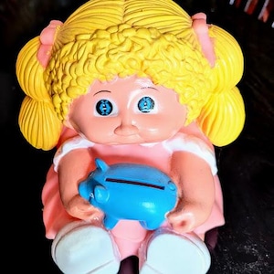 May include: A vintage doll figurine with yellow curly hair, blue eyes, and a pink dress. The doll is holding a blue piggy bank. The doll's feet are white. The figurine is a collectible item.